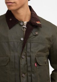 Ελαιόχρωμο μπουφάν με καφέ καπιτονέ γιακά, κλείσιμο με φερμουάρ και πολλαπλές μπροστινές τσέπες; διαθέτει μια μικρή κόκκινη ετικέτα με λογότυπο στην αριστερή πλευρά.