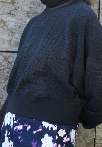 Černá mikina se strukturovaným krokodýlím vzorem, žebrovaným lemem a dlouhými rukávy. V kombinaci se kalhotami s květinovým vzorem v tmavých barvách.