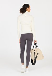 Suéter blanco de cuello alto acanalado, leggings grises y un bolso de duffel beige con correas negras. La modelo está de espaldas, mostrando el atuendo.