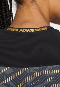 Svart atletisk skjorte med ribbet tekstur, med "VENUM PERFORMANCE" i gullskrift ved kragen og et mønstrete grått og gulldesign nedenfor.