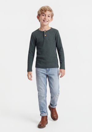 Lächelnder Junge mit blondem Haar trägt ein dunkles Langarm-Henley-Shirt, hellblaue Jeans und braune Stiefel und läuft vor einem schlichten Hintergrund nach vorne.