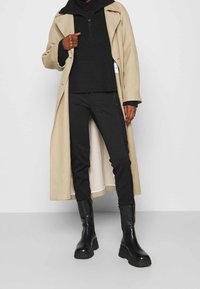 Beige lång kappa, svart ribbad tröja, svarta byxor och kraftiga svarta stövlar. Slät textur, lager-på-lager stil och minimalistisk design.