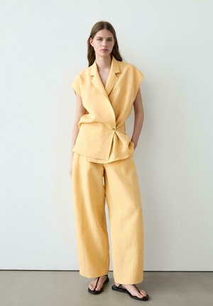 Femme portant un blazer sans manches jaune ample et un pantalon large assorti, debout contre un mur blanc uni, les mains dans les poches.