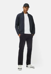 Donkerblauwe gewatteerde jas met een staande kraag, witte t-shirt eronder, zwarte corduroy broek en witte sneakers met marineblauwe accenten.