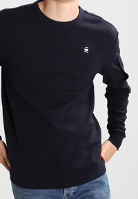 Mann trägt ein schlichtes schwarzes Langarmshirt mit einem kleinen weißen Logo auf der Brust und blaue Jeans, die Hände auf den Hüften.
