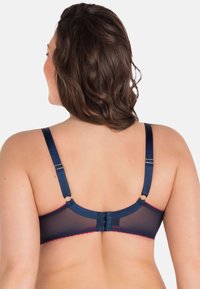 Sujeto azul marino con detalles de espalda transparente, tirantes ajustables y cierre de gancho y ojo. Presenta acentos rojos a lo largo de los bordes.