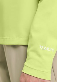 Limettengrünes Langarmhemd aus strukturiertem Stoff. Mit weißem "TEXADRI"-Logo am Bündchen. Nahtdetails sind am Ärmelrand sichtbar.