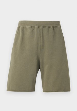 Άνδρας που στέκεται με τα χέρια στις τσέπες του ελιάς πράσινου σορτς, φοράει μαύρο κοντομάνικο πόλο, λευκές κάλτσες και καφέ δερμάτινα παπούτσια σε γκρι φόντο.