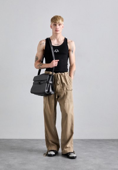 Tânăr cu tatuaje, îmbrăcat cu un top fără mâneci negru și pantaloni cargo bej largi, ținând o geantă de umăr din piele neagră, stând pe un podea gri.