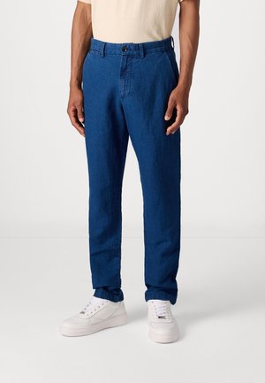 Mand iført blå bukser med lige pasform, hvide sneakers og en beige kortærmet skjorte, stående mod en ensfarvet baggrund.