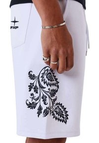 Main portant des bagues en argent et un bracelet repose contre un short blanc avec une broderie florale noire et le logo PXP près de la poche.