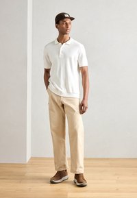 Cămașă polo albă cu mâneci scurte, pantaloni bej și adidași maro. Cămașa are un guler și nasturi, creând un aspect casual.