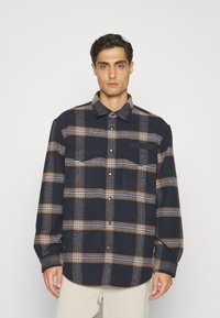 Camicia in flanella a quadri blu navy e marrone con chiusura a bottoni, due tasche sul petto e una texture morbida. Presenta un colletto e orlo arrotondato.