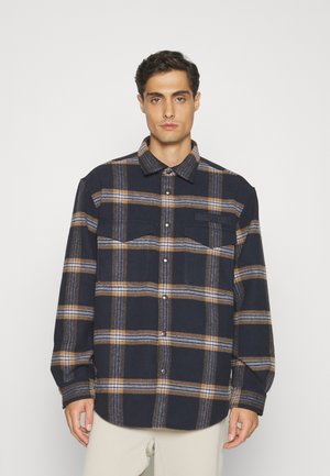 Camicia in flanella a quadri blu navy e marrone con chiusura a bottoni, due tasche sul petto e una texture morbida. Presenta un colletto e orlo arrotondato.