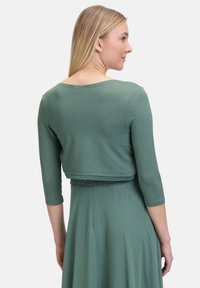 Zielony krótki top z trzema ćwierć rękawami, wykonany z miękkiego materiału, z okrągłym dekoltem i koronkowym wykończeniem u dołu.