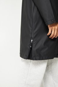 Black waterproof jacket features a smooth, matte finish, side zippers, and a relaxed fit, paired with gray sweatpants for contrast.