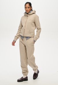 Felpa beige con zip e tasche frontali, abbinata a pantaloni della tuta coordinati. Camicia a righe visibile sotto, completata da mocassini scuri.