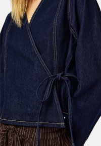 Haut portefeuille en denim bleu foncé avec manches larges, présentant des coutures contrastées et une ceinture nouée, porté avec un pantalon marron à rayures verticales.