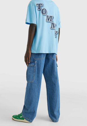 Vaaleansininen puuvillapaita, jossa on selässä rohkeasti painettu "TOMMY" tummilla kirjaimilla. Yhdistetty löysiin sinisiin farkkuasusteisiin.