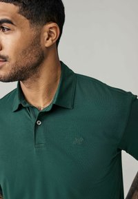 Grøn polo skjorte med tekstureret stof, to-knaps lukning og broderet logo på venstre bryst. Korte ærmer og klassisk kravedesign.