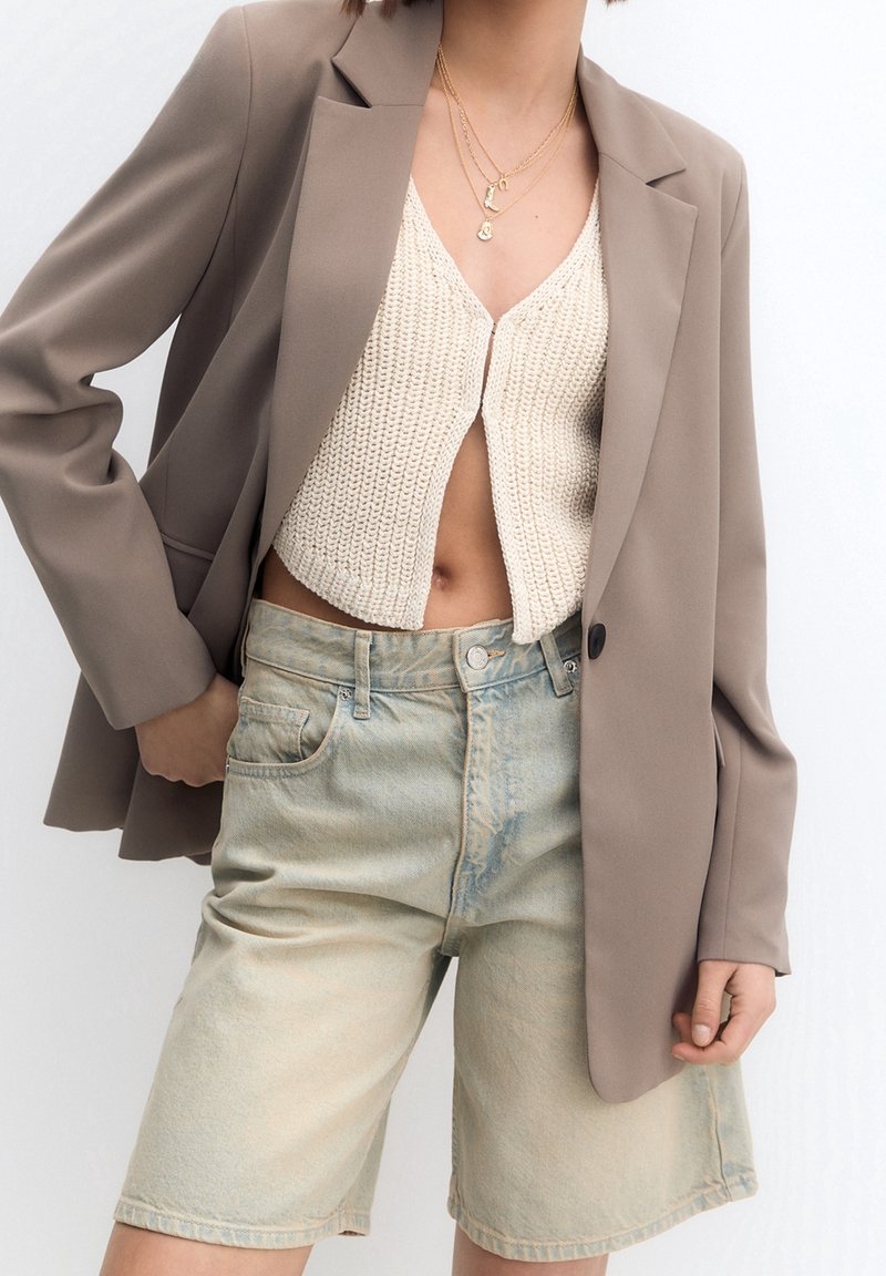 Beige blazer over en cremefarvet strikket cropped top, kombineret med lyseblå denimshorts. Blazeren har en enkel knap; toppen har en ribbet tekstur.