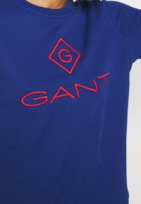 Modrý bavlněný svetr s červeně vyšitým textem a logem "GANT" ve tvaru diamantu na přední straně. Hladká textura, design s kulatým výstřihem.