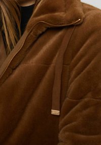 Gros plan d'une personne portant une veste en fausse fourrure marron douce avec une fermeture éclair et un cordon, montrant une partie de longs cheveux bruns et un haut noir.