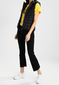 Czarna pikowana kamizelka z kapturem, żółta koszulka, czarne spódnice dzwonowe i białe buty wsuwane. Gładkie tekstury i nowoczesny krój.