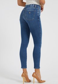 Mujer que lleva puestos unos pantalones skinny de tiro alto azules con el logo de Guess en el bolsillo trasero y zapatos de tacón alto color beige, de pie contra un fondo liso.