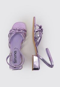 Sandalias moradas de tacón con material sintético brillante, punta abierta, correas entrelazadas, un pequeño tacón cuadrado y un lazo decorativo en la correa frontal.