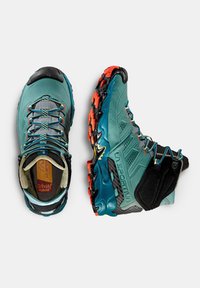 Scarpe da trail running in turchese e nero, con una tomaia testurizzata, accenti arancioni sulla suola e un sistema di allacciatura sicuro per una maggiore stabilità.