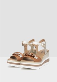 Brown wedge sandals with a metallic gold strap, featuring a chain accent, a textured soft footbed, and layered white and brown sole.