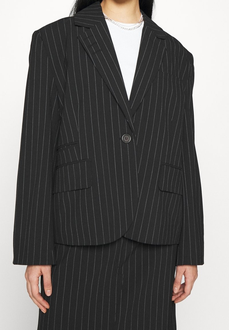 Femme portant un blazer noir à fines rayures avec un seul bouton, poches à rabat, assorti à une jupe rayée et un collier chaîne argenté sur une chemise blanche.