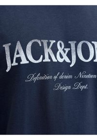 Tissu bleu foncé avec texte blanc "JACK & JONES" en grande police et texte cursif plus petit "Definition of denim Nineteen Design Dept."