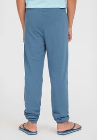 Blå bomullssweatpants med elastiska mudd och midja, slät textur. Bärs med blå flip-flops, sedd bakifrån.