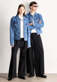 Világoskék farmerdzsekik gombos részletekkel, fehér pólók fölött viselve. A szetteket fekete, széles szárú nadrágok egészítik ki. Simogató textúrák és szabott illeszkedés.