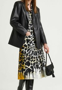 Blazer in ecopelle nera su un vestito fantasia con stampa giraffa in nero e giallo. Accessoriato con una piccola borsa nera con frange.