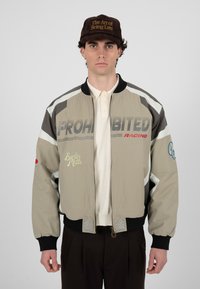 Jeune homme portant une veste de course beige et noire avec le texte « PROHIBITED », un polo crème, un pantalon foncé et une casquette marron portant l'inscription « The Art of Being Late ».