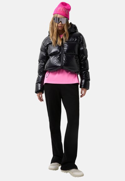 Kvinde iført en skinnende sort dunjakke, en lyserød hue og T-shirt, reflekterende solbriller, sorte bukser og hvide sneakers står på hvid baggrund.
