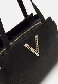 Svart handväska i texturerat läder, med ett guldzipper och en framträdande guldig V-formad detalj på framsidan.