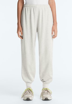 Világosszürke melegítőnadrág rugalmas derékkal és szegéllyel, világos sportcipőkkel, melyeken sárgászöld fűzők láthatók, deréktől lábig mutatva.