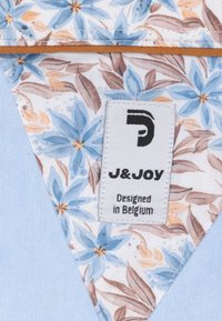 Bloemstoffen patch met blauwe en bruine bladeren, omzoomd met oranje stiksel, bevestigd aan een blauwe textuurstof. Etiket leest "J&Joy, Ontworpen in België."