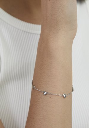 Silver bracelet featuring small heart-shaped charms linked by a delicate chain, resting on a skin-toned wrist.