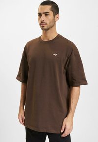 Brun oversized t-skjorte laget av myk bomull, med rund hals og korte ermer, med en liten hvit logo på brystet.