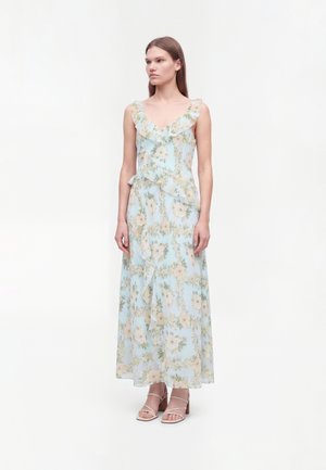 Mujer con vestido maxi sin mangas azul claro con estampado floral y volantes, de pie frente a un fondo blanco liso, lleva tacones beige con tiras.