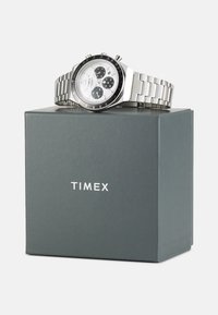 Silberne Edelstahl-Uhr mit einem weißen Zifferblatt und schwarzen Akzenten, ausgestattet mit Chronographen-Subdials, auf einer grauen Timex-Box platziert.