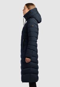 Marineblå pufferjakke med høj krave, lynlåslommer og en quilted design. Har en snoretræk i hætten og en tætsiddende silhuet.
