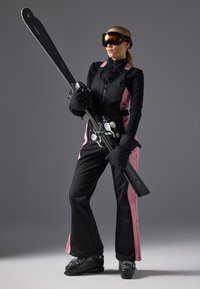 Skioutfit in zwart en roze met een getailleerd ontwerp, handschoenen en skibril. Houdt zwarte skies vast met witte bindingen. Sneeuwlaarzen zichtbaar.