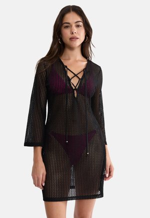 Kobieta ubrana w czarną, siateczkową sukienkę z długim rękawem na ramiączkach, nałożoną na ciemnofioletowy bikini z krzyżującymi się wiązaniami na piersiach, stojąca na białym tle.