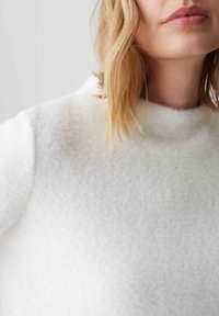 Maglione bianco e morbido con un colletto alto, caratterizzato da una consistenza soffice e una vestibilità leggermente ampia. Il dettaglio mostra parte del collo e della spalla.